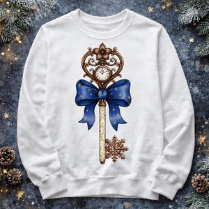 May include: White sweatshirt featuring a detailed illustration of a key with a clock face, a blue bow with gold stars, and a snowflake. The key is brown and gold, with a decorative floral design at the top.