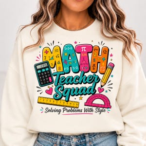 May include: A cream-colored sweatshirt featuring a colorful graphic with the text "MATH Teacher Squad" and "Solving Problems With Style." The design includes a calculator, a yellow pencil, a yellow ruler, a pink protractor, and math symbols.
