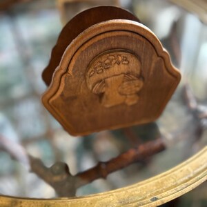 May include: A wooden napkin holder with a carved mushroom design. The holder is a light brown color and has a scalloped edge. The mushroom design is centered on the front of the holder.