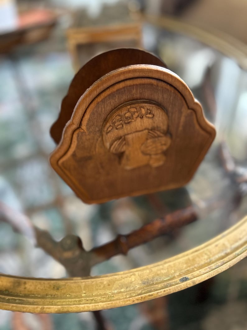 May include: A wooden napkin holder with a carved mushroom design. The holder is a light brown color and has a scalloped edge. The mushroom design is centered on the front of the holder.