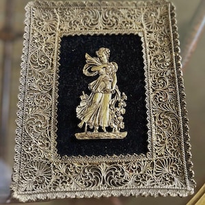 May include: A gold-toned filigree frame with a black velvet interior. A gold-colored figure of a woman holding a vase and flowers is centered on the velvet. The frame has an ornate, scrolling design.