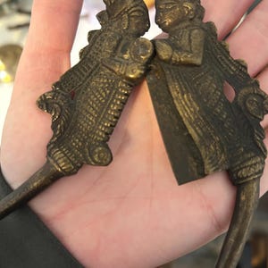 May include: Two antique bronze-colored metal spoons. Each spoon features a detailed figure of a person in traditional attire. The figures face each other, with intricate designs on their clothing and headwear. The spoons have long, slender handles.