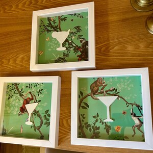 May include: Three white picture frames with green and white patterned paper inside. Each frame features a white paper cutout of a martini glass. The frames are hanging on a wood wall.
