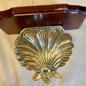 May include: A decorative wall shelf featuring a dark brown wooden top and a gold-colored, seashell-shaped bracket. The bracket has a detailed, textured design. The shelf is designed to be mounted on a wall and is suitable for displaying small items.