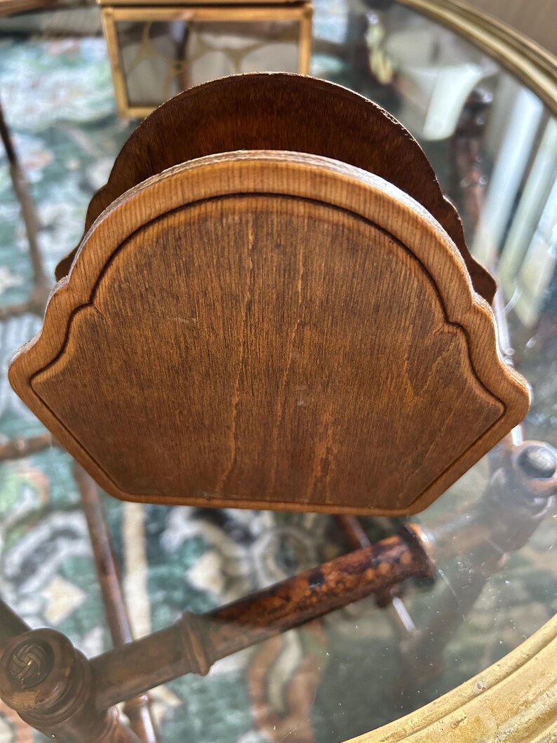 May include: A wooden napkin holder with a scalloped edge design. The holder is made of a medium-toned wood with visible grain, giving it a natural and rustic appearance. The holder is designed to hold napkins and is likely for use on a dining table or countertop.