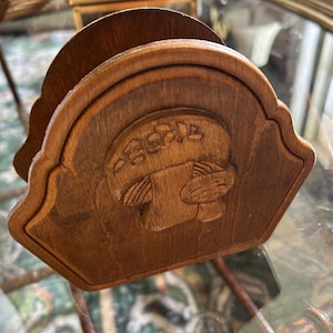 May include: A wooden napkin holder with a carved design. The holder is a medium brown color and features a decorative, arched top. The carved design appears to be a stylized image of a person or animal. The holder is sitting on a glass surface.