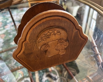 May include: A wooden napkin holder with a carved design. The holder is a medium brown color and features a decorative, arched top. The carved design appears to be a stylized image of a person or animal. The holder is sitting on a glass surface.
