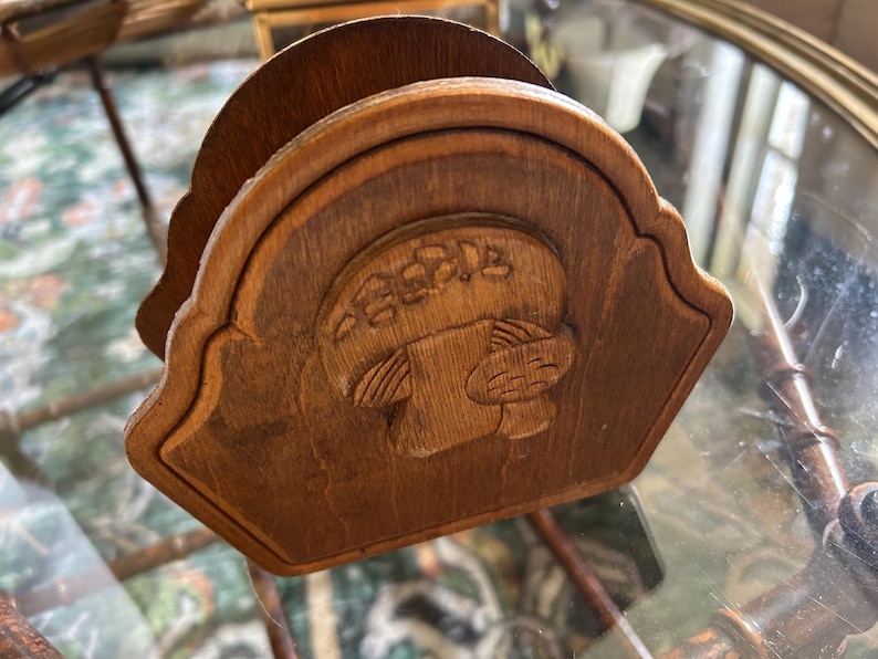 May include: A wooden napkin holder with a carved design. The holder is a medium brown color and features a decorative, arched top. The carved design appears to be a stylized image of a person or animal. The holder is sitting on a glass surface.