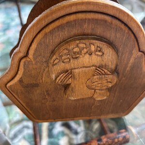 May include: Wooden napkin holder with a carved mushroom design. The holder is made of a light brown wood and has a curved top. The mushroom is in the center of the holder and has a detailed design.