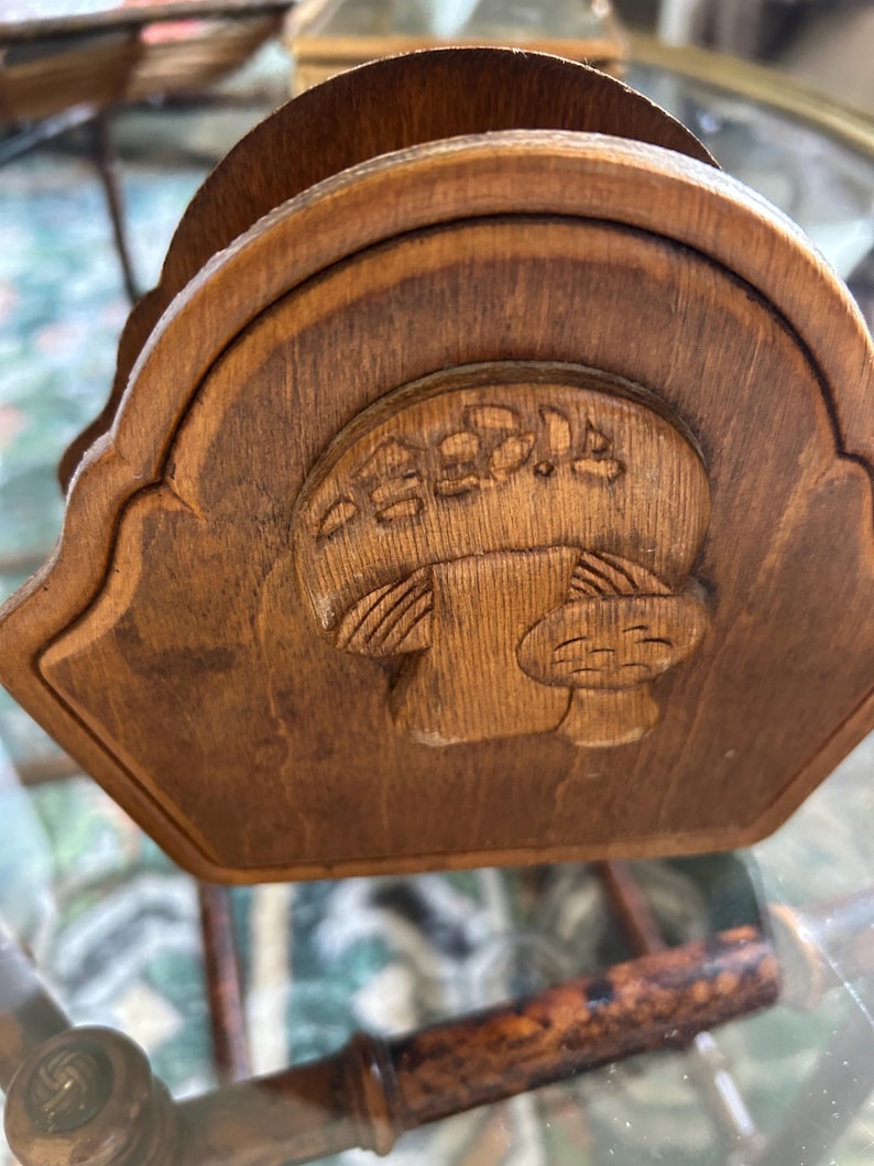 May include: Wooden napkin holder with a carved mushroom design. The holder is made of a light brown wood and has a curved top. The mushroom is in the center of the holder and has a detailed design.