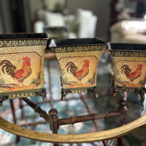 May include: Set of three decorative metal planters with a rooster design. The planters have a distressed, vintage look with a gold and green color scheme. Each planter features a rooster illustration and sits on decorative feet.