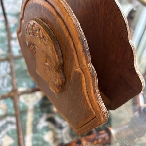 May include: A wooden napkin holder with a carved relief of a classical figure. The holder is made of dark brown wood and has a curved design. The relief is on the front panel, and the holder is designed to hold napkins.