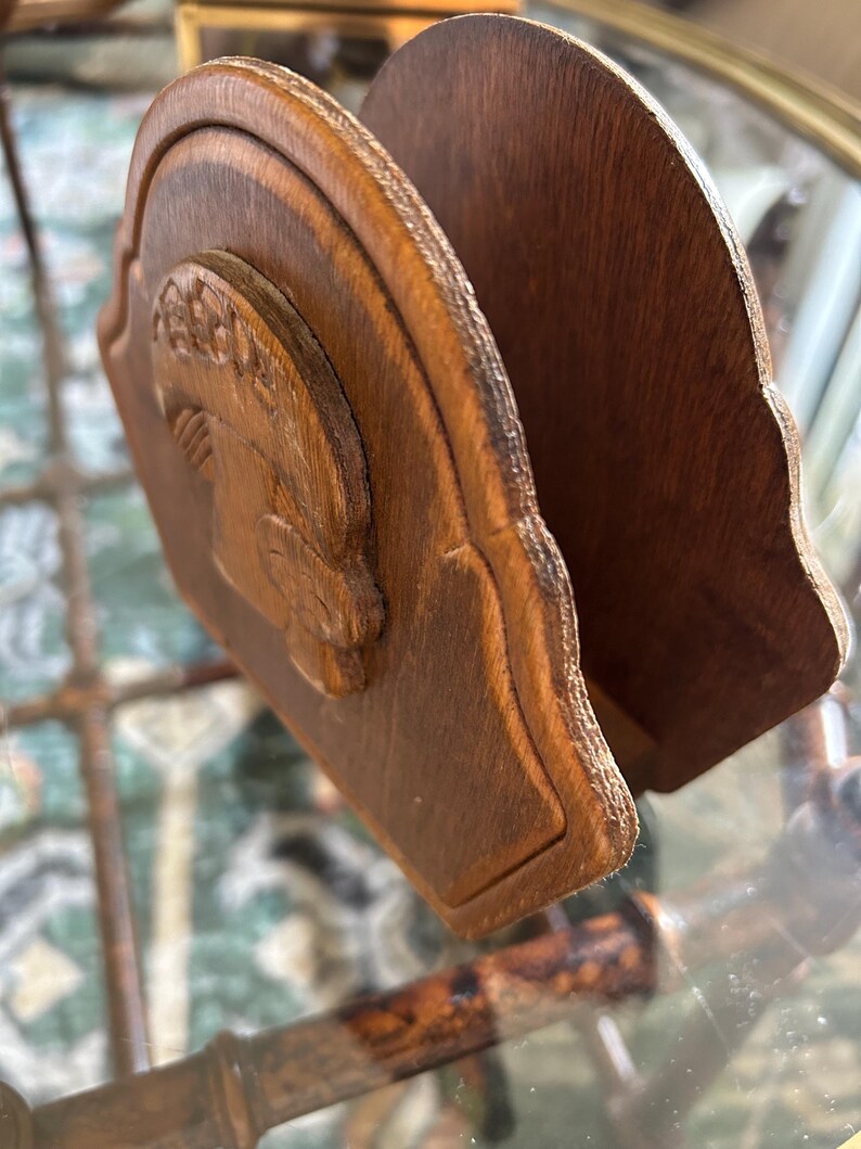 May include: A wooden napkin holder with a carved relief of a classical figure. The holder is made of dark brown wood and has a curved design. The relief is on the front panel, and the holder is designed to hold napkins.