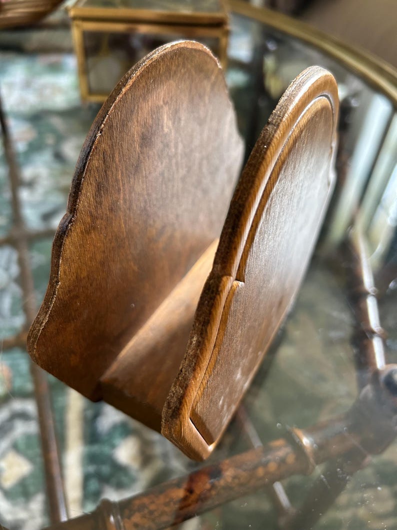 May include: A wooden napkin holder with a scalloped edge design. The holder is made of light brown wood and has a slot to hold napkins. The holder is angled, showing the interior space. The wood grain is visible.