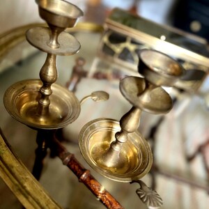 May include: Two antique brass candle holders with a classic design. Each holder features a round base, a central stem, and a small cup at the top for holding a candle. The holders have a warm, golden hue and are placed on a reflective surface.