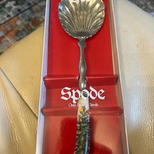 May include: A Spode Christmas Tree serving spoon in its original box. The spoon features a silver-colored, shell-shaped head and a white handle decorated with a Christmas tree design. The box has the words "Spode Christmas Tree" printed on it.