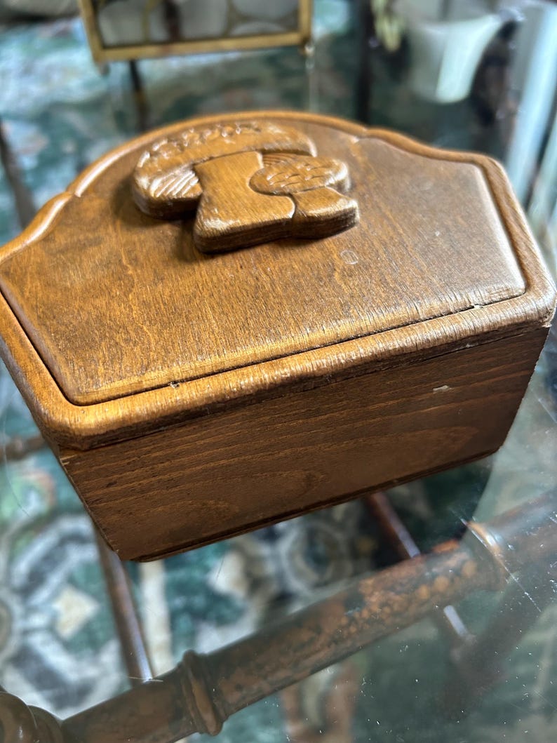 May include: A small, rectangular wooden box with a carved mushroom design on the lid. The box is stained a warm brown color and appears to be vintage. The lid has a curved top edge.