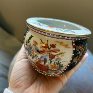 May include: A small, round ceramic bowl with a wide opening. The bowl features a cream-colored base with painted birds and floral designs in red, blue, and yellow. The rim is trimmed with gold and a dark red border. The sides have a black and green pattern.