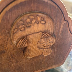 May include: A wooden decorative piece with a carved mushroom design. The mushroom has a smiling face and Japanese text above it. The wood has a warm, brown tone, and the carving is detailed.