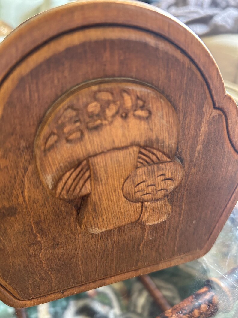 May include: A wooden decorative piece with a carved mushroom design. The mushroom has a smiling face and Japanese text above it. The wood has a warm, brown tone, and the carving is detailed.