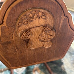 May include: A wooden decorative piece with a carved mushroom design. The mushroom has a smiling face and is surrounded by carved details. The wood has a warm, brown tone, and the piece has a curved top edge.