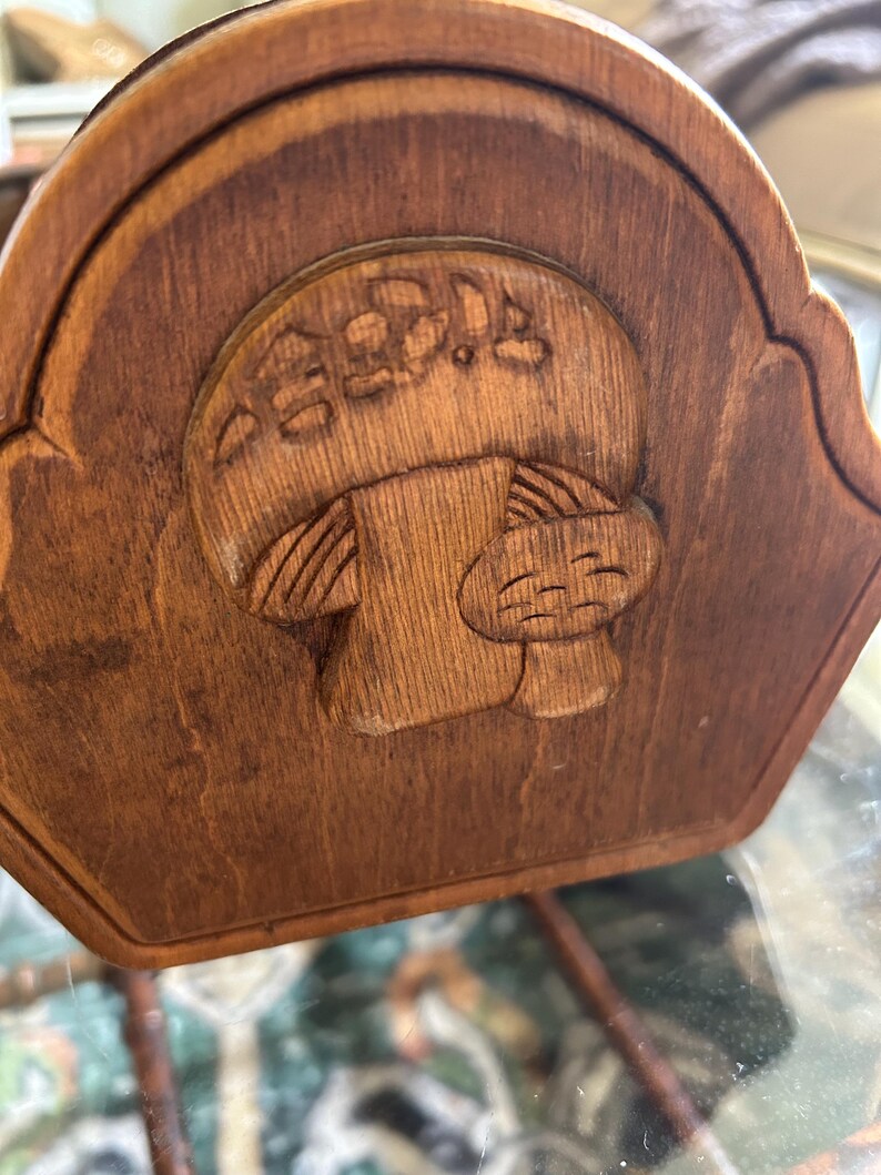 May include: A wooden decorative piece with a carved mushroom design. The mushroom has a smiling face and is surrounded by carved details. The wood has a warm, brown tone, and the piece has a curved top edge.