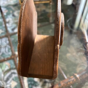 May include: A wooden napkin holder with a curved, scalloped design. The holder is made of light brown wood and has a rectangular base. The sides are curved and feature a decorative edge. The holder is designed to hold napkins upright.