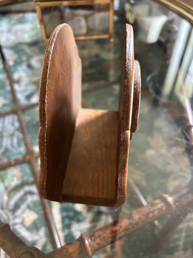 May include: A wooden napkin holder with a curved, scalloped design. The holder is made of light brown wood and has a rectangular base. The sides are curved and feature a decorative edge. The holder is designed to hold napkins upright.