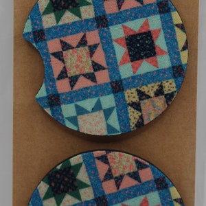 May include: Two round, fabric-covered car coasters with a patchwork design featuring blue, pink, green, and black stars. The coasters are packaged on a brown card with the text "Car Coasters Handmade with love".