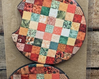 Puede incluir: Dos posavasos para coche con un diseño de patchwork en tonos rojos, naranjas, verdes y beige. Los posavasos son redondos con un recorte curvo. El embalaje dice "Car Coasters Handmade with love."