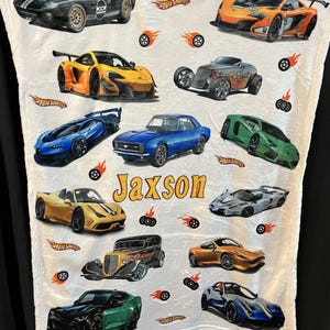 May include: A white fleece blanket with a Hot Wheels theme. The blanket features a variety of colorful cars, including a blue and white car, a yellow and black car, a red and black car, and a green and black car. The blanket also features the Hot Wheels logo and the name "Jaxson".