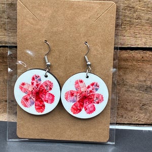 May include: A pair of round earrings with a floral design. Each earring has a white background with a red and pink flower pattern. The earrings are displayed on a brown card with silver ear wires.