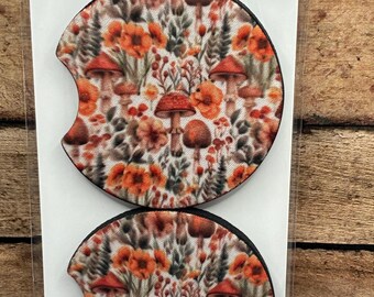 May include: Two car coasters with a colorful floral and mushroom design. The coasters have a black border and a cutout for easy removal. The packaging reads "Car Coaster" and states the coasters help keep cup holders clean.