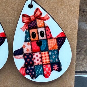 Puede incluir: Un par de pendientes con tem&aacute;tica de Halloween. Cada pendiente presenta una forma de l&aacute;grima blanca con un dise&ntilde;o de fantasma sonriente. El fantasma est&aacute; hecho de cuadrados de patchwork coloridos y tiene un lazo rojo en la cabeza.