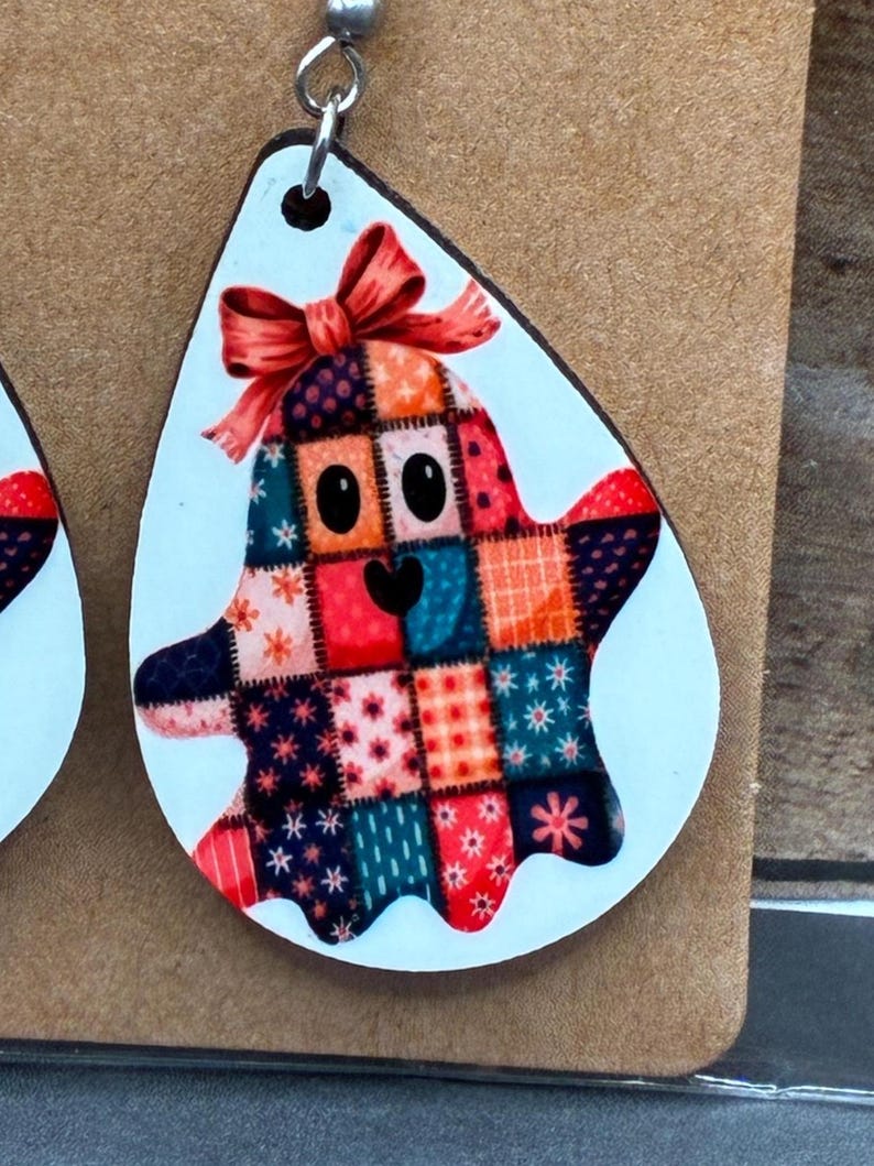 Puede incluir: Un par de pendientes con tem&aacute;tica de Halloween. Cada pendiente presenta una forma de l&aacute;grima blanca con un dise&ntilde;o de fantasma sonriente. El fantasma est&aacute; hecho de cuadrados de patchwork coloridos y tiene un lazo rojo en la cabeza.