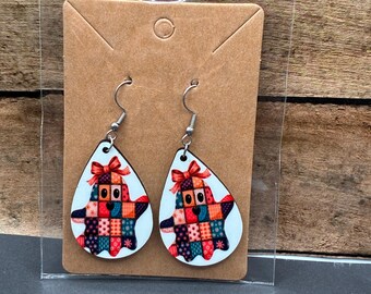 May include: A pair of teardrop-shaped earrings featuring a patchwork ghost design. The ghosts have a red bow and are in a clear plastic package on a brown card. The earrings have silver-colored fish hook ear wires.