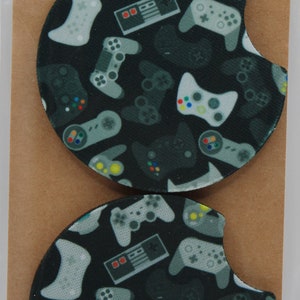 May include: Two round car coasters with a black background and a pattern of gray and white video game controllers. The coasters are handmade and say "Car Coasters Handmade with love" on the packaging.