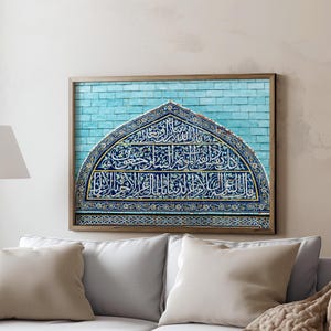 May include: A framed artwork featuring a blue and white arched design with intricate calligraphy. The artwork is mounted on a light blue brick background. The frame is a light brown color. The artwork hangs above a white sofa with beige pillows.