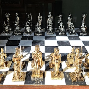 Könnte beinhalten: Ein komplettes Schachspiel mit goldenen und silbernen Figuren auf einem schwarz-weißen Schachbrett. Die goldenen Figuren befinden sich im Vordergrund, die silbernen im Hintergrund. Das Brett steht auf einem dunklen Holztisch.