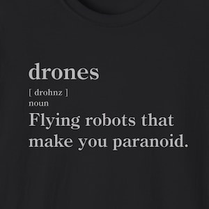 Op de afbeelding: Zwart T-shirt met ronde hals met de witte definitie van "drones" erop gedrukt. De definitie luidt: "Vliegende robots die je paranoïde maken."