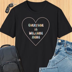 Może przedstawiać: Czarny T-shirt z sercowatym wzorem. Serce zawiera napis "Everyone is Welcome Here" kolorowymi literami.