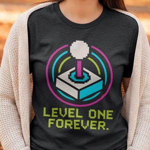 Könnte beinhalten: Dunkelgraues T-Shirt mit einem pixeligen Arcade-Joystick-Design. Das Design umfasst eine weiße Kugel, rosa, blaue und grüne Bögen sowie den Text "LEVEL ONE FOREVER". Das Design ist auf dem T-Shirt zentriert.