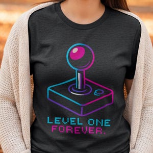 Op de afbeelding: Donkergrijs t-shirt met een retro arcade joystick afbeelding in blauw, paars en roze. De tekst "LEVEL ONE FOREVER" staat eronder. Een leuk, nostalgisch ontwerp voor gamers.