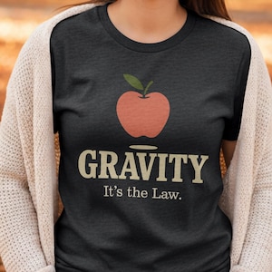 Könnte beinhalten: Dunkelgraues T-Shirt mit einer pfirsichfarbenen Apfelgrafik und einem grünen Blatt. Das Wort "GRAVITY" ist in beiger Schrift gedruckt, darunter der Satz "It's the Law."