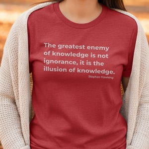 Puede incluir: Camiseta roja con la cita "El mayor enemigo del conocimiento no es la ignorancia, es la ilusión del conocimiento" de Stephen Hawking en blanco. Se lleva con una rebeca color crema.
