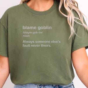 以下が含まれることがあります： オリーブグリーンのTシャツ。ライトグレーで「blame goblin」というフレーズがプリントされています。テキストには定義、発音、そして「Always someone else's fault never theirs.」というフレーズが含まれています。