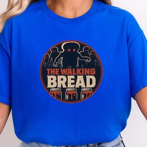 Puede incluir: Camiseta azul real con un gráfico circular. El gráfico presenta el texto "THE WALKING BREAD" y figuras con forma de pan con un diseño de zombi. Tres tarros de mermelada están en la parte inferior del gráfico.