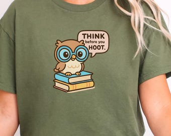 May include: Olive green t-shirt featuring a cartoon owl wearing blue glasses, perched on a stack of books. The owl has a speech bubble that says, "THINK before you HOOT."