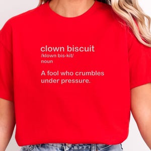以下が含まれることがあります： 「clown biscuit」という言葉と発音、定義が書かれた赤いTシャツ。定義は「プレッシャーに屈する愚か者」という意味。テキストはサンセリフ体で表示されています。