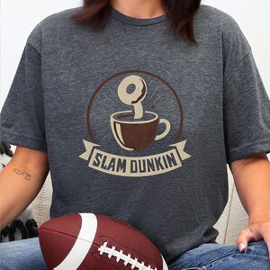 Peut inclure: T-shirt gris foncé avec un motif marron d'une tasse à café avec un beignet et le texte "SLAM DUNKIN" sur une bannière. Un ballon de football marron et blanc est au premier plan.